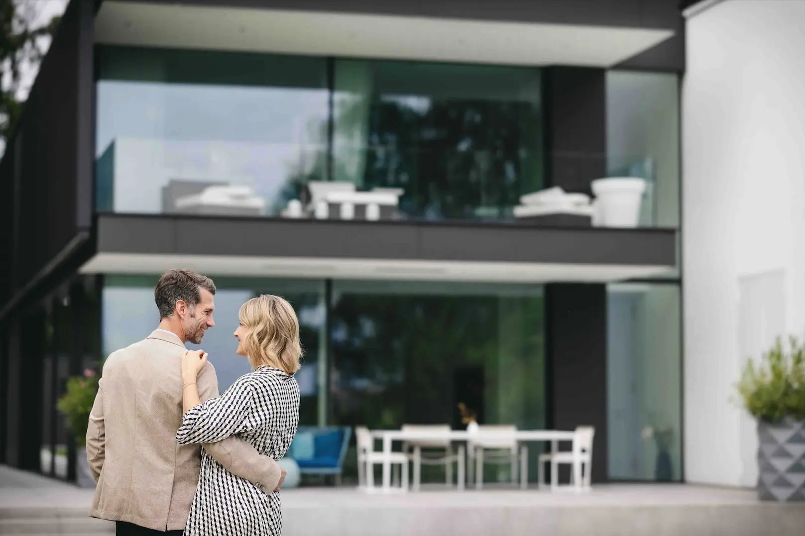 Glücklicher Paar vor moderner Villa mit Glasfront, Terrasse und Gartenmöbeln