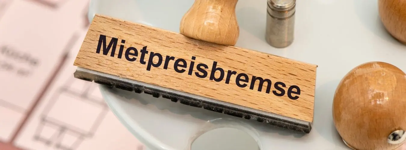 Ein Holzstempel ist beschriftet mit Mietpreisbremse über einem Grundriss einer Mietwohnung
