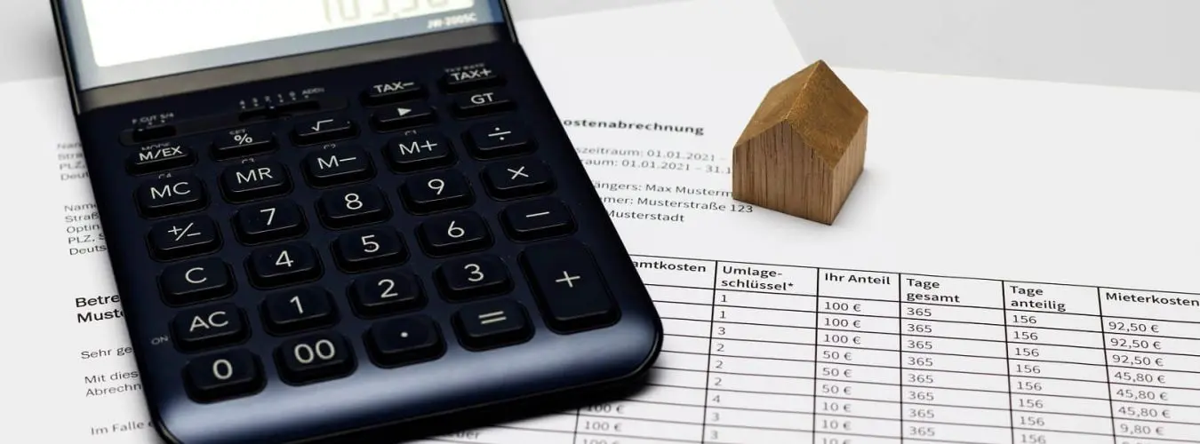 Taschenrechner mit Nebenkostenabrechnung und Modellhaus aus Holz auf einem Schreibtisch